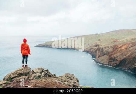 Trail runner femmina vestito con felpa con cappuccio arancione sportivo e cappuccio rosso poggiato sulla scogliera e godendo la vista sull'oceano Atlantico sulla penisola di Ponta de Sao Lourenço Foto Stock