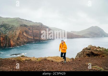 Trail runner uomo vestito arancione impermeabile giacca, running tights e scarpe jogging da Atlantic Ocean Bay gamma sulla penisola Ponta de Sao Lourenço -t Foto Stock