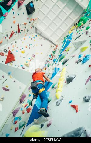 Ragazzo adolescente al muro di arrampicata al coperto sala. Ragazzo sta arrampicandosi usando una corda superiore e un'imbracatura per arrampicarsi e qualcuno che lo tardò dal pavimento. Adolescente attivo Foto Stock