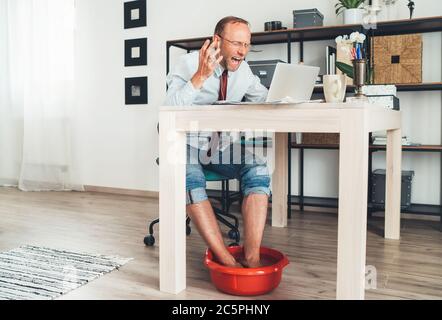 Situazione comica moderna dell'ufficio domestico. Uomo d'affari hanno video chiamata emotiva che chattano sul laptop e che si alza i piedi in bagno caldo piedi sotto table.Distanc Foto Stock