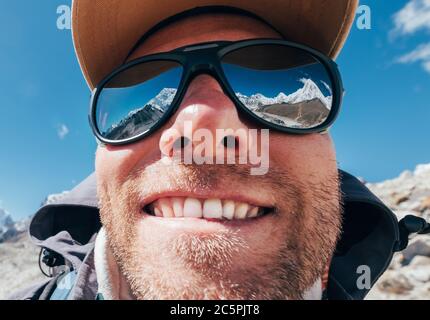 Obiettivo ultra-grandangolare ritratto scatto di alta quota montagna sorridente unshaven happy escursionista in cappellino da baseball con cime innevate e catena montuosa accanto a h. Foto Stock