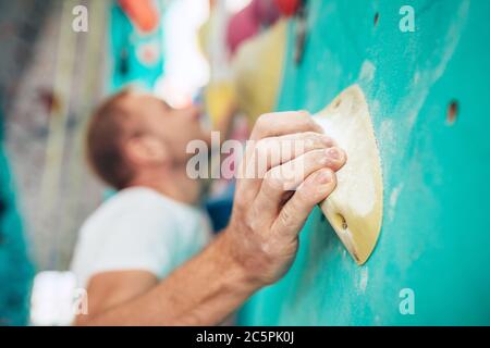 Primo piano di palma dell'uomo ricoperte di gesso (magnesio) che si aggrappano all'elemento di presa per arrampicata in interno mentre l'uomo sale. Gli scalatori non concentrati si affacciano sulla schiena Foto Stock