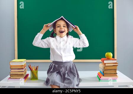 Ritorno a scuola. Una studentessa con un libro sopra la testa sorride sullo sfondo della lavagna della scuola. Foto Stock