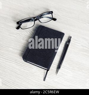 Foto di taccuino nero chiuso, bicchieri e matita su sfondo di tavola in legno chiaro. Foto Stock