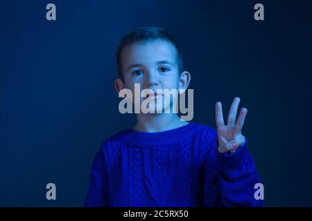 Ritratto in studio di una brunetta Boy che conta sulle dita, mostra tre dita, tonificanti in un classico colore blu. Foto Stock
