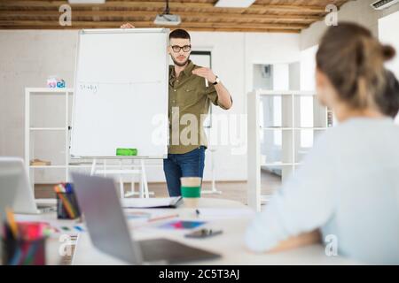 Giovane uomo d'affari in occhiali e camicia in piedi vicino a lavagna bianca mentre discutono attentamente nuovo progetto con i colleghi. Gruppo di creativi Foto Stock