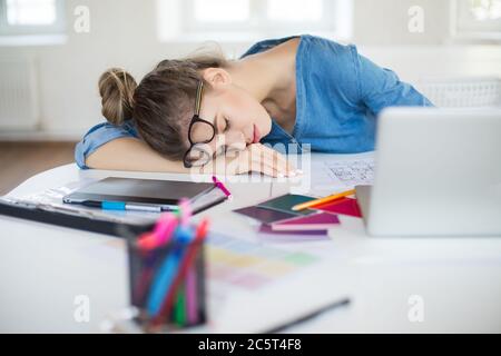 Donna giovane stanca con occhiali sulla testa che dormono sulla scrivania con il computer portatile vicino mentre si passa il tempo in un ufficio moderno Foto Stock