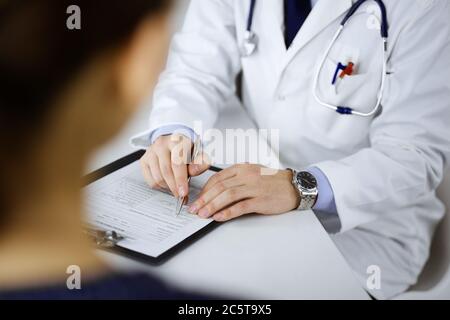 Un medico sta parlando con il suo paziente, mentre si siede insieme alla scrivania nel gabinetto in un ospedale. Medico che usa gli appunti per il riempimento Foto Stock