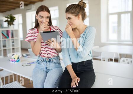 Sorridenti ragazze sedute sulla scrivania con tablet che girano felicemente insieme a un ufficio vuoto sullo sfondo Foto Stock