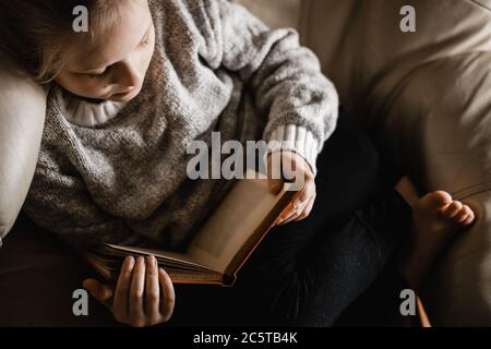 Una ragazza caucasica bionda seduta arricciata su una sedia che legge un libro Foto Stock