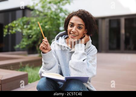 Bella ragazza con capelli ricci scuri seduta con libro aperto sulle ginocchia e felicemente guardando in macchina fotografica mentre studiano nel cortile dell'università Foto Stock