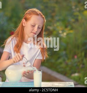 Il latte fresco versa da una caraffa in un bicchiere una ragazza dai capelli rossi. La luce solare calda illumina la ragazza e il latte sul tavolo. Vetro di latte si leva su un wo Foto Stock