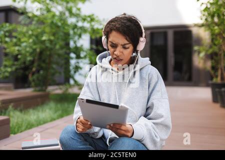 Giovane donna sconvolta in cuffie seduta e guardando stupito in tablet mentre si passa il tempo nel cortile dell'università Foto Stock