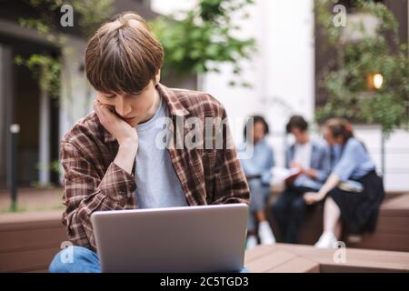 Foto di un giovane stanco seduto sulla panchina con un laptop in ginocchio e pensando mentre si passa il tempo nel cortile dell'università con gli studenti sullo sfondo Foto Stock