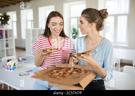 Due giovani sorridenti seduti sulla scrivania che parlano e mangiano volentieri la pizza mentre si passa il tempo insieme in un ufficio moderno Foto Stock