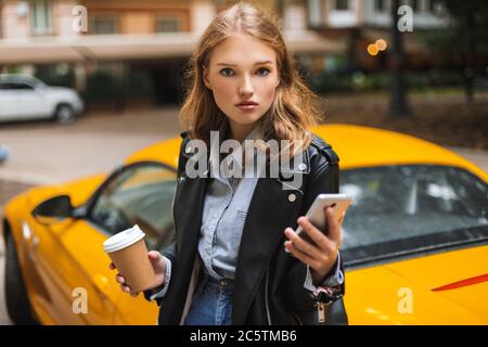 Giovane bella donna in pelle giacca che tiene la tazza di caffè per andare e cellulare in mani mentre guardando pensieroso in macchina fotografica con auto sportiva gialla Foto Stock