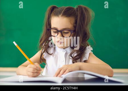 Concetto di ritorno a scuola. Una ragazza intelligente in occhiali scrive con una matita in un notebook mentre si siede a un tavolo in classe. Foto Stock