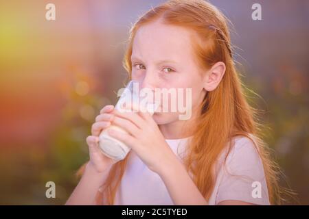 La ragazza dai capelli rossi beve latte fresco da un bicchiere sullo sfondo di una bella mattina soleggiata nel villaggio. Bere latte è molto utile per l'hea Foto Stock