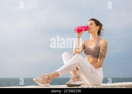 Giovane bella donna con top sportivo e leggings bianchi che tengono una bottiglia sportiva rosa e acqua potabile con vista mare su sfondo Foto Stock