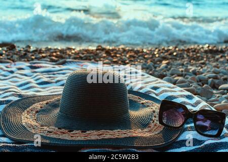 Concetto di vacanza estiva con cappello di paglia, telo da spiaggia e occhiali da sole su una spiaggia di ghiaia.oceano blu in background alla luce del sole. Foto Stock