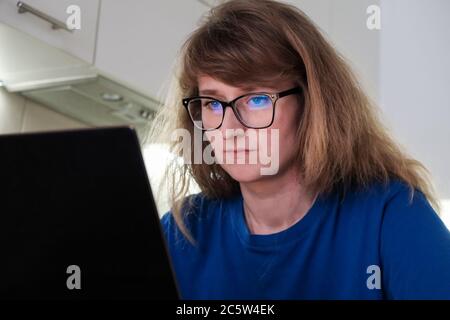 Donna giovane concentrata in occhiali guarda un monitor laptop mentre si siede a casa Foto Stock