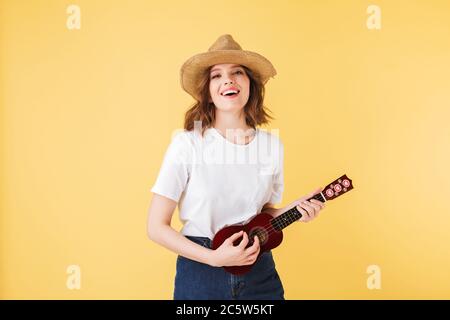 Ritratto di bella donna sorridente in cappello di paglia in piedi con la piccola chitarra e suonando su di esso mentre felicemente guardando in macchina fotografica su sfondo rosa. Foto Stock