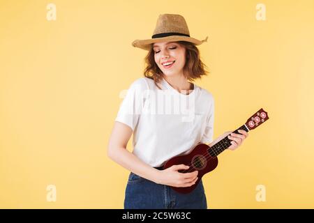 Ritratto di donna sorridente in cappello di paglia in piedi con la piccola chitarra e giocante felicemente su di esso su sfondo rosa Foto Stock