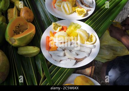 Frutta esotica tropicale servita sulla spiaggia da un uomo locale. Lastre di carta bianca sulle foglie di palma verdi. Isola delle Seychelles, Praslin. Vista dall'alto. Tro Foto Stock