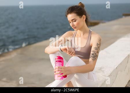 Giovane donna pensierosa con top sportivo e leggings bianchi che tengono in mano una bottiglia sportiva rosa mentre si guarda sognatamente a mano con vista mare Foto Stock