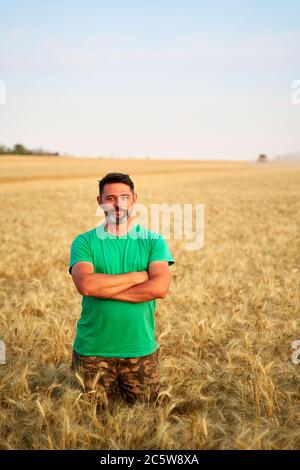 Agricoltore felice in piedi orgogliosamente in campo di grano con le braccia incrociate sul petto. Agronomo che indossa l'uniforme aziendale, guardando la macchina fotografica su terreni agricoli. Ricco Foto Stock