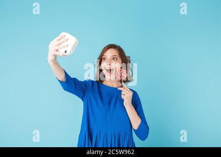 Ritratto di giovane donna in abito in piedi con lollipop e scattando una foto carina sulla sua piccola macchina fotografica su sfondo blu Foto Stock