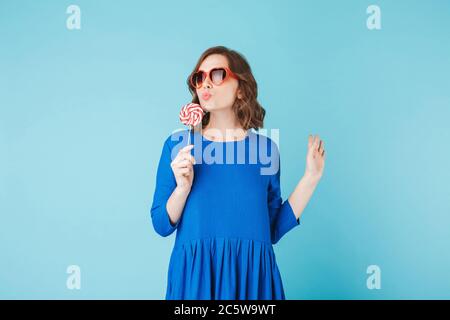 Foto di bella signora in occhiali da sole cuore e vestito in piedi con caramella lollipop in mano su sfondo blu Foto Stock