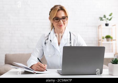 Il paziente parla con il medico utilizzando la videochiamata sul computer portatile. Donna in camice bianco prendere appunti in notebook Foto Stock