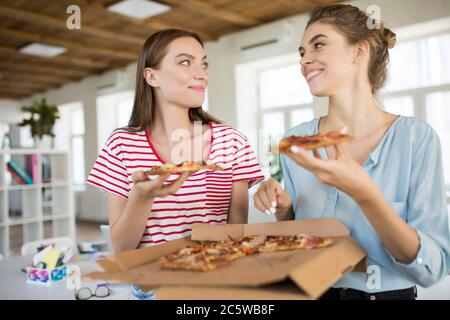 Due giovani sorridenti seduti sulla scrivania con pizza che si guardano a vicenda mentre si passa il tempo insieme in un ufficio moderno Foto Stock