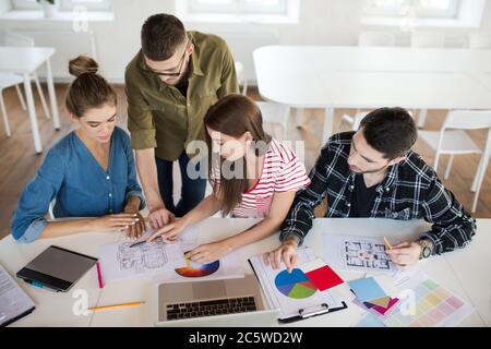 Gruppo di uomini e donne d'affari che lavorano con schizzi e computer portatile mentre si passa il tempo al lavoro. Giovani creativi discutono attentamente del lavoro Foto Stock