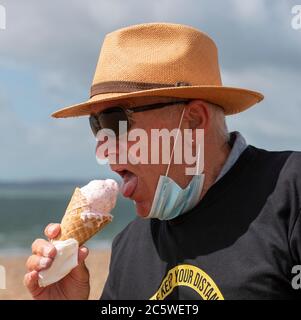 Southsea, Portsmouth, Inghilterra, Regno Unito. 2020. Uomo anziano sulla spiaggia che indossa la maschera, mangiare gelato e tenere la t-shirt distanza durante il Covid-19 ep Foto Stock