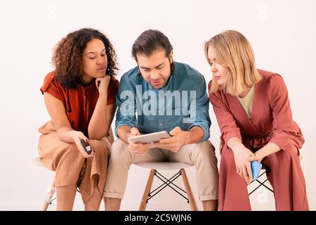 Due giovani donne in casualwear che guardano lo schermo del touchpad tenuto da un ragazzo mentre guardano film online o video di social network Foto Stock