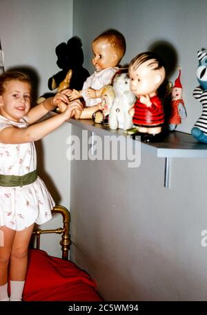 Giovane bambina di 4 anni in camera da letto con ottone Letto che mostra la sua collezione di bambole giocattolo, incluso il personaggio Linus Dal cartone animato Peanuts negli Stati Uniti negli anni '60 Foto Stock