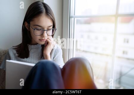 Carino libro di lettura della ragazza adolescente a casa mentre si siede sulla soglia della finestra Foto Stock