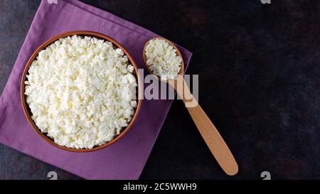 Ricotta il formaggio e cucchiaio di legno su sfondo scuro. Formaggio fresco casolare in una ciotola. Piatto di argilla con formaggio morbido su un tovagliolo di lino. Vista dall'alto. Copia sp Foto Stock