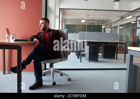 Ritratto a lunghezza intera di un uomo d'affari elegante seduto sul posto di lavoro e guardando via mentre si lavora in un ufficio vuoto contemporaneo, spazio di copia Foto Stock