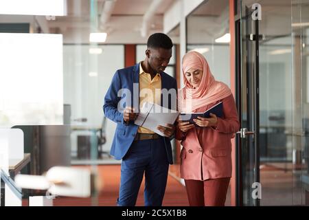 Ritratto di afroamericano che parla con giovane uomo d'affari musulmano e che tiene documenti mentre cammina verso la macchina fotografica nell'interno dell'ufficio, copia spa Foto Stock