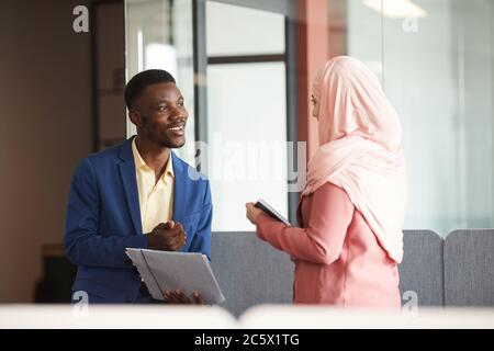 Vista laterale ritratto di sorridente afroamericano che parla con giovane donna d'affari musulmana e che tiene documenti in ufficio moderno interno, copia spazio Foto Stock