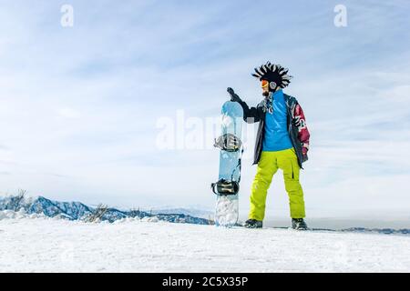 Snowboarder con brandello in maschera da sci con occhiali e un cappello mohawk in pelliccia grande sullo sfondo di un cielo e montagne innevate invernali Foto Stock