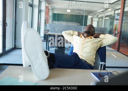 Ritratto di un uomo d'affari afroamericano contemporaneo che si rilassa sul posto di lavoro in ufficio con i piedi sulla scrivania, spazio per la copia Foto Stock