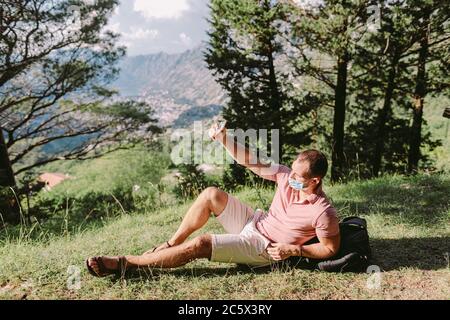 uomo che viaggia indossando una maschera protettiva per il viso e utilizzando uno smartphone in montagna Foto Stock