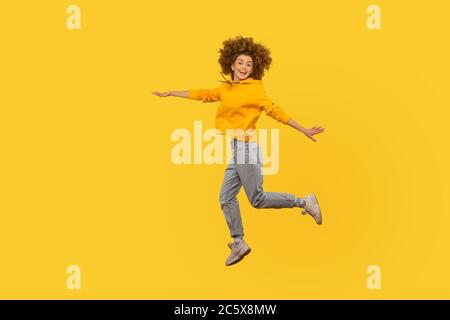 Ritratto di entusiasta vivace ed energica ragazza ricci-capelli in stile urbano felpa con cappuccio e jeans che volano in aria, godendo di vita felice, sentimento inspirato Foto Stock