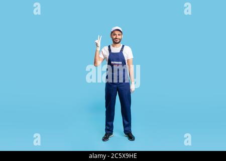 Uomo di riparazione esperto a tutta lunghezza in tute e guanti protettivi in piedi, mostrando gesto di vittoria e sorridendo soddisfatto. Professione di servizio in Foto Stock