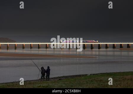 2 treni a cremagliera della classe 153 attraversano il viadotto di Arnside sulla linea ferroviaria della costa di Cumbria con pescatori che pescano e un cielo nero tempestoso Foto Stock