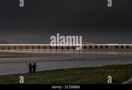 2 treni a cremagliera della classe 153 attraversano il viadotto di Arnside sulla linea ferroviaria della costa di Cumbria con pescatori che pescano e un cielo nero tempestoso Foto Stock